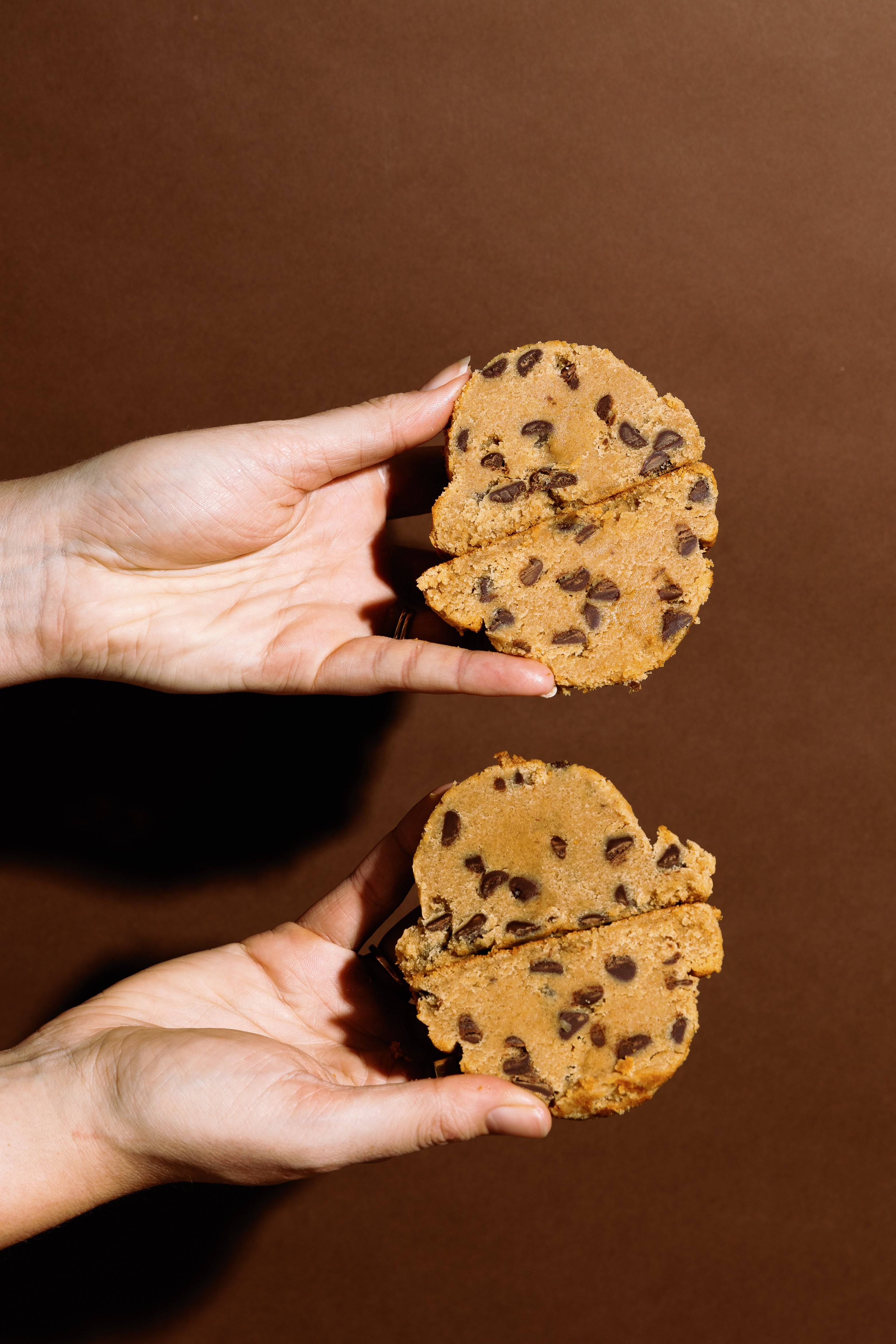 Two hands holding up half-pound cookies, showing the dense and delicious inside, filled with chocolate chips